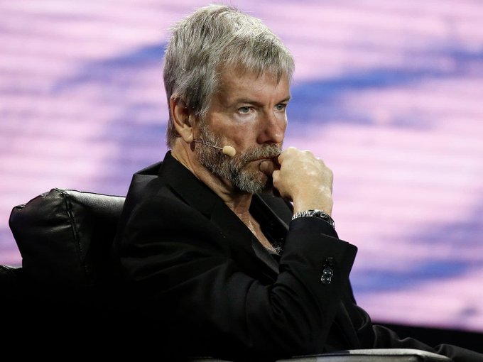 A man with gray hair and a beard, wearing a black jacket and a headset microphone, sits thoughtfully with his hand on his chin, likely at a conference or speaking event. The image appears alongside news of Michael Saylor's Strategy selling 1,637 BTC worth over $102 million, creating a notable contrast with his widely cited "Never sell your Bitcoin" philosophy.