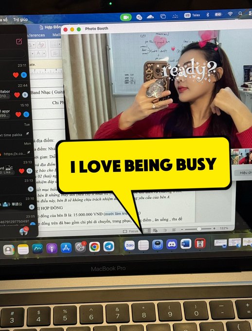 A MacBook screen showing a Photo Booth preview of a woman taking a selfie with 'ready?' text and a yellow speech bubble reading 'I LOVE BEING BUSY' overlaid on the image.