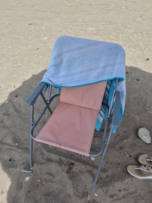 A pink folding beach chair sits on sand with a blue towel draped over its backrest, accompanied by a pair of sandals nearby.