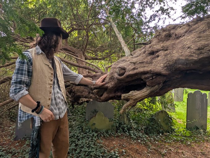 A person wearing a brown hat, plaid shirt, and beige vest standing in a wooded area with old gravestones, touching a large gnarled tree trunk or root formation.