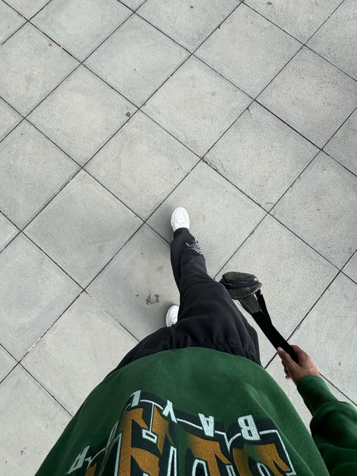 A first-person perspective photo looking down at feet in white sneakers and black pants walking on grey tiled pavement, holding a camera strap, with a green hoodie featuring a graphic in the foreground.