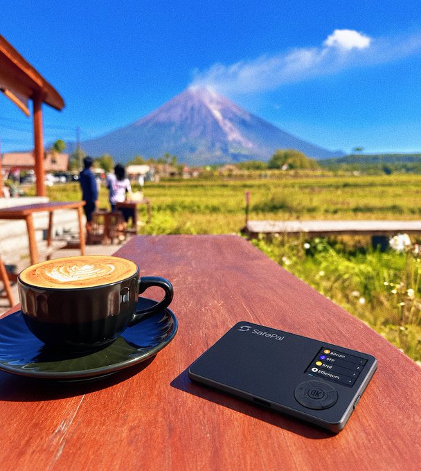 A latte with latte art sits on a wooden table beside a SafePal hardware wallet displaying Bitcoin, ETH, and Ethereum options, with a volcano and rice fields in the background—capturing the essence of "Weekend check: who's watching charts?" as even scenic coffee breaks call for a quick crypto portfolio glance.