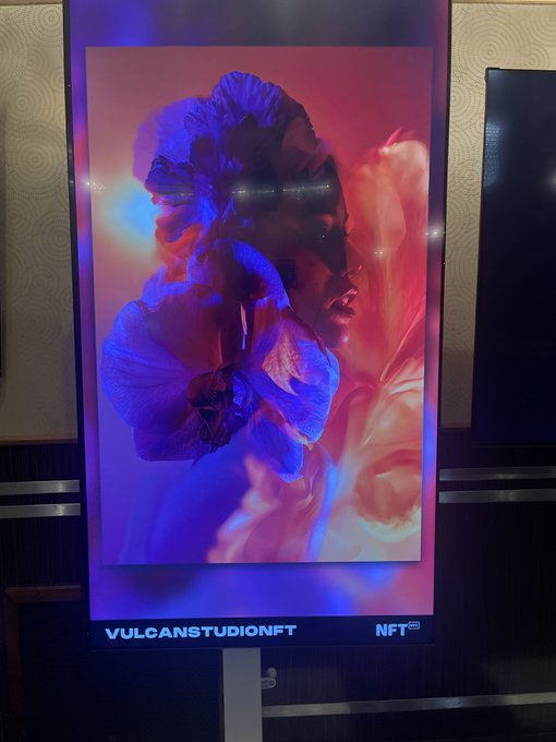 A vertical digital NFT display screen showing an artistic portrait of a woman with flowing hair rendered in blue and red neon tones, labeled 'VULCANSTUDIONFT NFT' at the bottom.