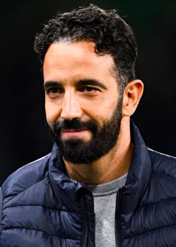 A close-up portrait of a man with dark curly hair and a full beard, wearing a dark blue puffer jacket over a grey shirt, smiling slightly.