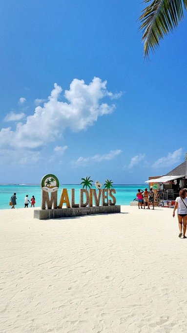 Tropical beach scene featuring a large 'MALDIVES' sign on white sand, turquoise water, a clear blue sky with clouds, and tourists walking nearby under a palm tree.
