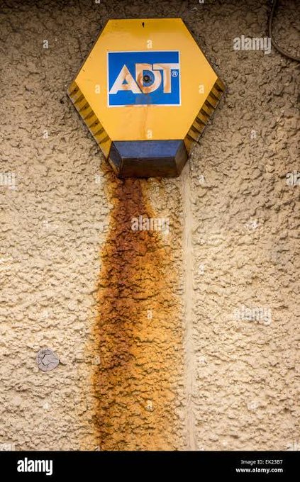 A yellow hexagonal ADT security alarm box mounted on a textured stucco wall, with a prominent rust stain dripping down from its base.