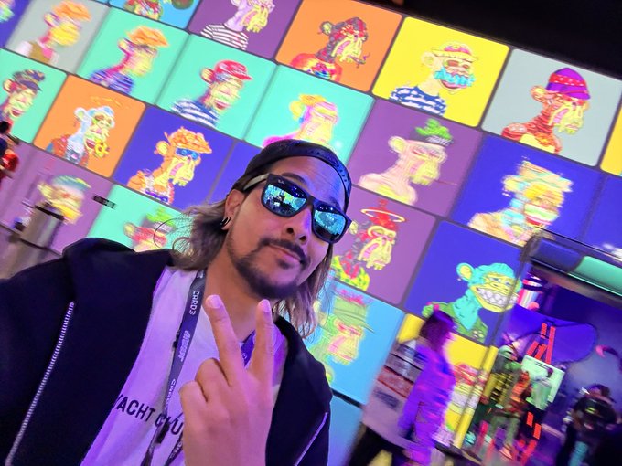 A man with long hair, sunglasses, and a black cap takes a selfie flashing a peace sign in front of a vibrant display of colorful Bored Ape Yacht Club NFT artwork, with "YACHT CL..." visible on his shirt, signing off with "gn fam."