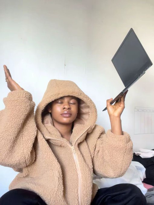 A person wearing a tan sherpa fleece hoodie poses with eyes closed, holding a laptop up in one hand while raising the other in a relaxed gesture.