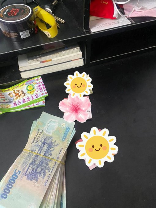 Vietnamese currency (50,000 dong notes) bundled with rubber bands sits on a black desk next to magazines, a lottery ticket, and an energy drink can, decorated with sun and hibiscus flower stickers.
