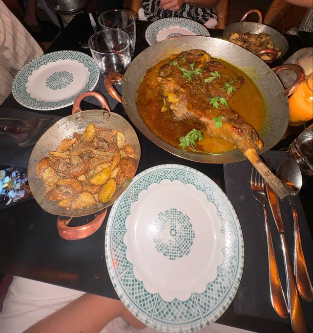 A restaurant table featuring a large lamb shank dish in a copper pan, accompanied by seasoned potato wedges in a smaller copper pan, with decorative white and teal patterned plates and glassware.