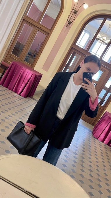 A woman taking a mirror selfie in an ornate room with pink tablecloths, wearing a black blazer with pink cuffs over a white t-shirt.