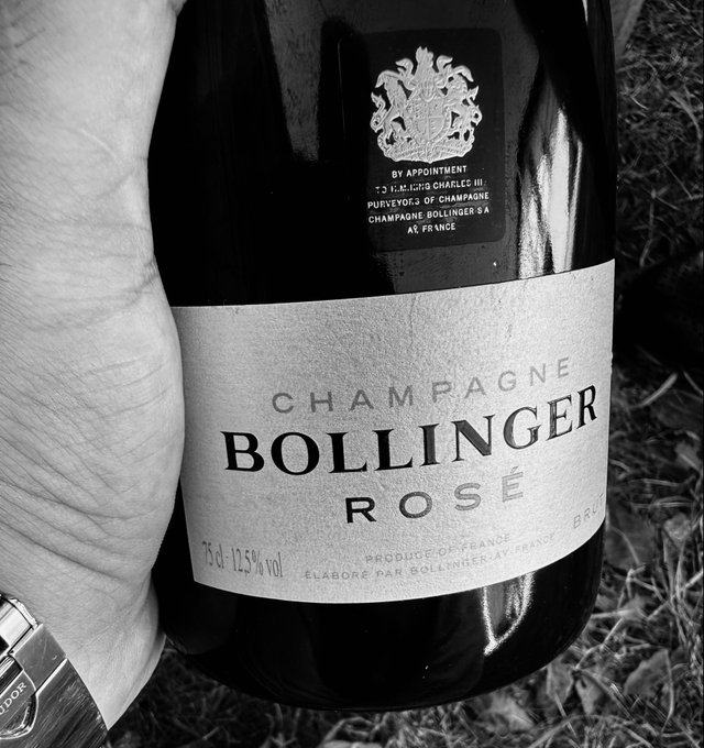 A hand holds a bottle of Champagne Bollinger Rosé (75cl, 12.5% vol., produced in France) against a backdrop of dry grass, suggesting a weekend celebration of taking a break from trading to enjoy the moment rather than chasing further gains—even if the price dumps.