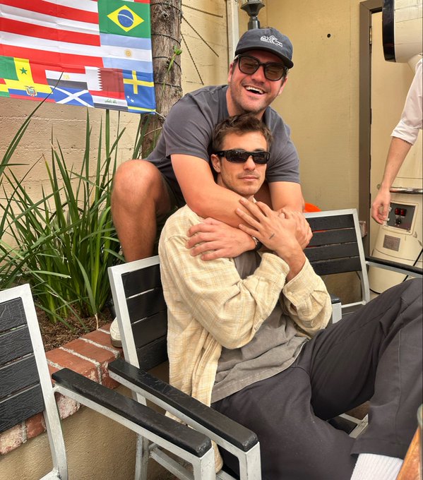 Two men pose closely together outdoors on a patio with black and white chairs, with various international flags including Brazil, Argentina, Senegal, and Scotland visible in the background.