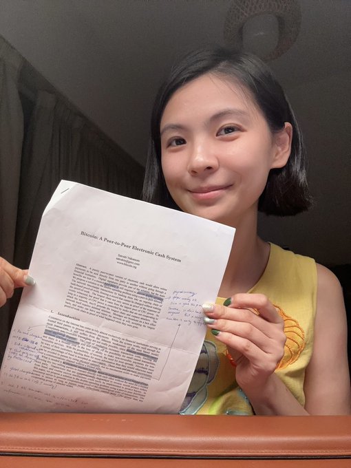 A smiling young woman in a yellow shirt holds up the original 'Bitcoin: A Peer-to-Peer Electronic Cash System' whitepaper by Satoshi Nakamoto, which is covered in handwritten annotations.