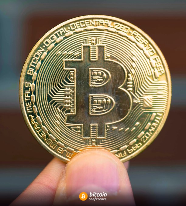A close-up shows a hand holding a gold-colored physical Bitcoin commemorative coin engraved with "BITCOIN DIGITAL DECENTRALIZED PEER TO PEER," "VIRES IN NUMERIS," and "FINE COPPER," perfectly illustrating the tangible representation of holding Bitcoin as discussed in the post.
