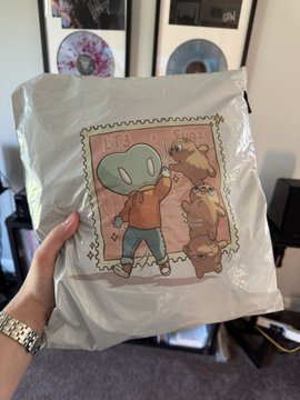 A person's hand holds up a packaged, light gray tote bag with an illustration of an alien in an orange sweater and two teddy bears under the text 'LET'S PLAY SHOT.'