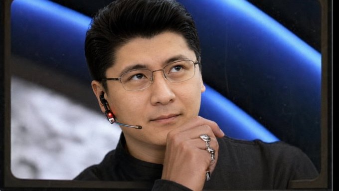 A man wearing glasses and a black shirt with an earpiece microphone rests his hand near his chin in a thoughtful pose, likely appearing as a panelist or speaker at a conference. The contemplative expression paired with the satirical post about broken promises of utility updates suggests someone appearing reflective amid criticism for failing to deliver on crypto project commitments.