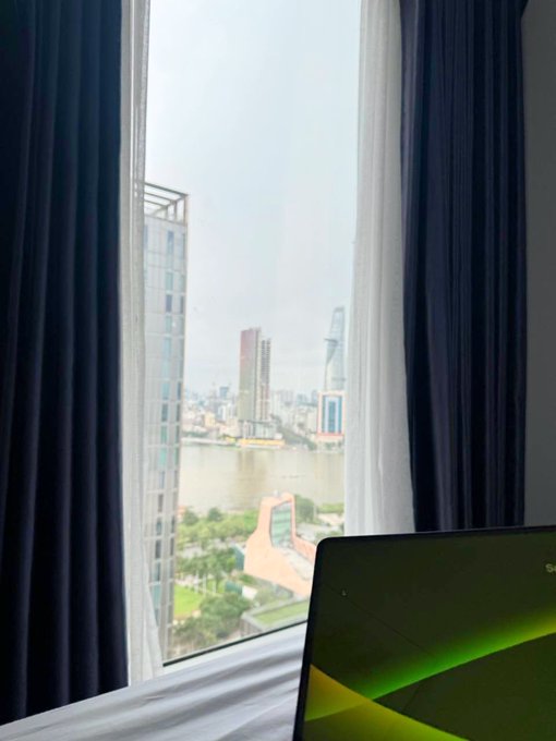A view through a partially curtained window shows a cityscape with a river and tall buildings, with the edge of a laptop visible in the foreground.