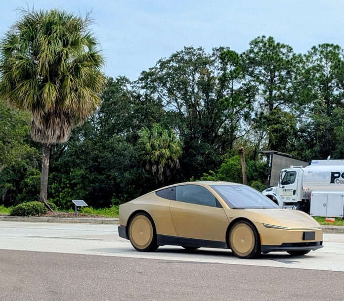 A tan/gold-colored Tesla Cybertruck is parked on pavement with palm trees and a white utility truck in the background.