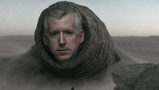 A dramatic portrait of a man with a serious expression emerging from a rocky, hood-like formation in a barren landscape, with a tiny silhouetted figure in the distance.