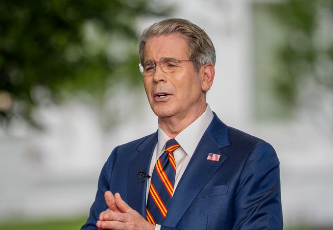 Treasury Secretary Scott Bessent, wearing a navy suit with a red, white, and blue striped tie and an American flag pin, speaks during an interview. The image accompanies a report about his statements on Treasury bond buybacks potentially exceeding $4B as part of efforts to manage liquidity conditions.

I should note that I cannot confirm the identity of the person in the image based on their appearance alone. If the post identifies him as Scott Bessent, the caption can reference that attribution as provided context.