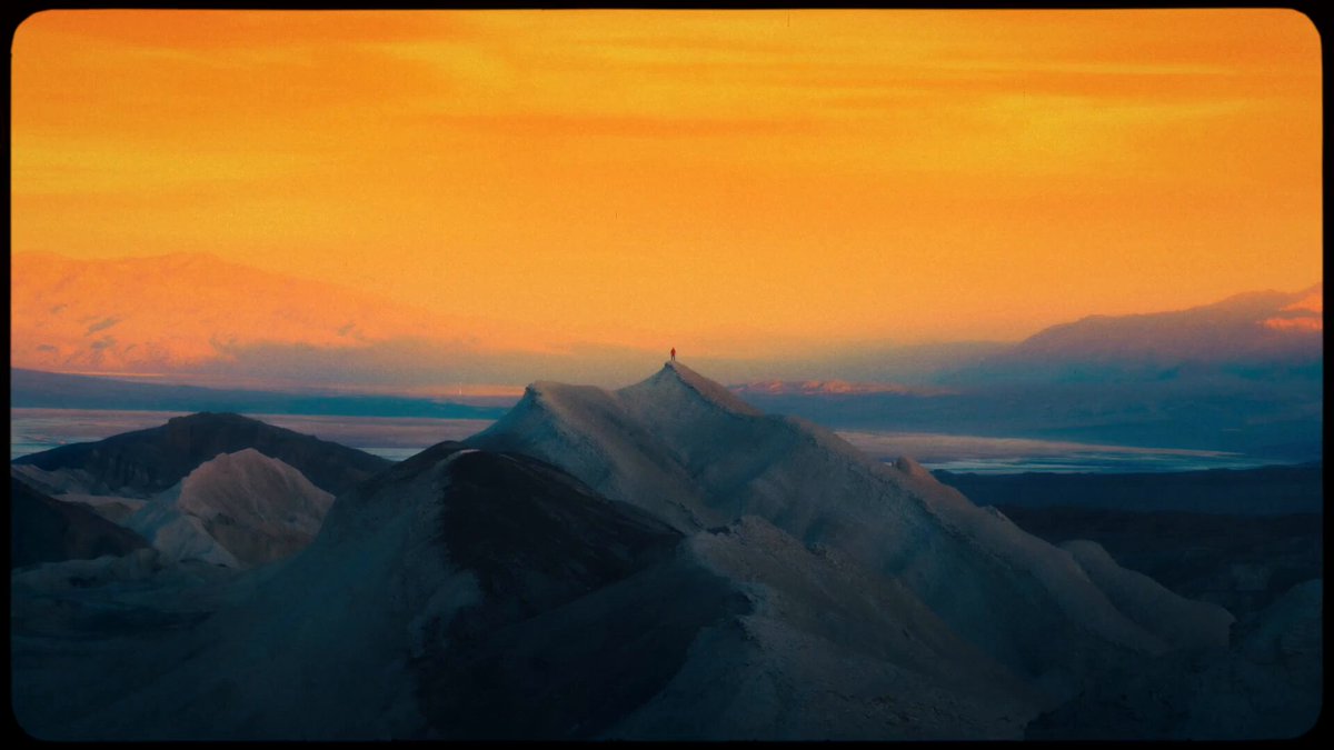 The image features a breathtaking view of a mountain range with a sunset casting an orange glow over the landscape. The sky transitions from a deep blue at the horizon to a warm orange near the mountains, creating a stunning backdrop for the scene. In the foreground, there is a person standing on top of one of the peaks, adding a sense of scale and grandeur to the image.