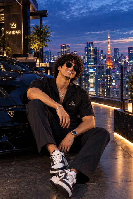 Man with curly hair in sunglasses sitting on the ground next to a black Lamborghini, with Tokyo Tower and the city skyline in the background.