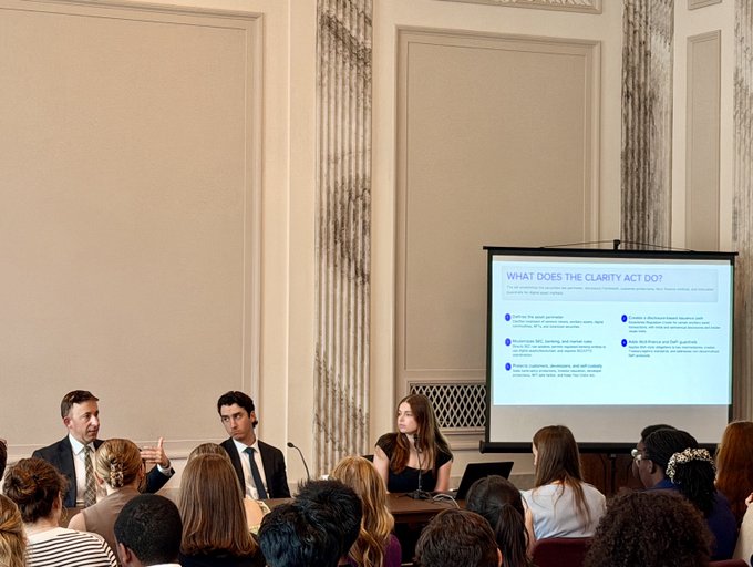 A panel discussion in an elegant room with attendees, featuring speakers presenting on a slide titled 'WHAT DOES THE CLARITY ACT DO?' displayed on a projector screen.