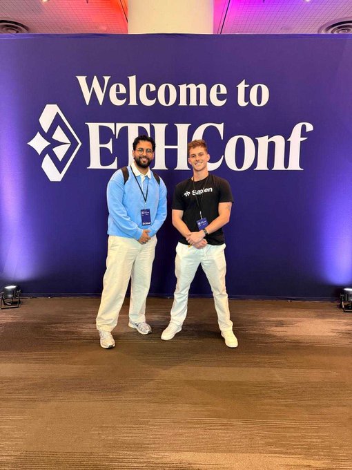 The image features two men standing next to a large purple banner that says "Welcome to EthCon". The men are wearing backpacks, indicating they might be attending an event or conference related to ethical artificial intelligence (AI). They appear to be posing for a photo in front of the banner, showcasing their participation and interest in the event.
