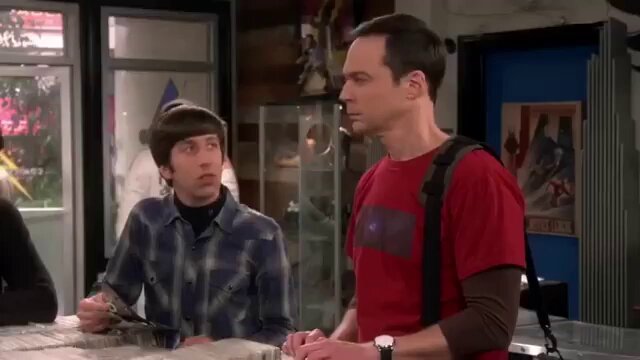 The image shows two men sitting at a table, with one man wearing a red shirt and the other wearing a blue plaid shirt. They are both looking at something off-camera, possibly engaged in conversation or observing an object nearby. The background features a window that provides natural light to the scene.