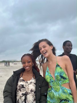 A casual photo of two smiling women standing together on a beach with cloudy skies, with a man standing in the background.