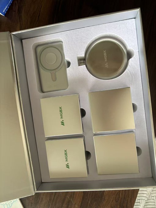 An unboxed MGBX promotional gift set containing a beige thermos/cup and four matching square product boxes arranged in a presentation tray.