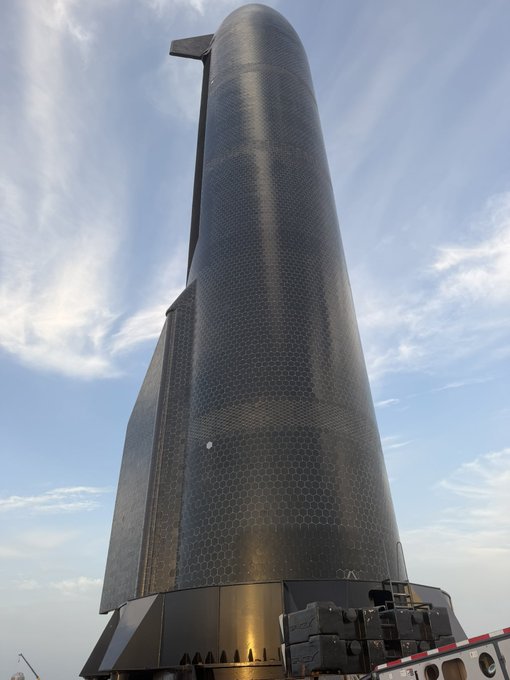A towering black rocket with a hexagonal tile pattern stands vertically against a partly cloudy sky, appearing fully assembled and ready for launch or transport. The image represents the "Now" phase, showcasing the completed spacecraft after its transformation from earlier stages.