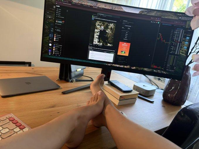 A person rests their bare feet on a wooden desk while working at a computer with multiple windows open, with the caption joking that this casual, feet-up setup is typical behavior for the average $TOESCOIN holder, suggesting a laid-back attitude toward their crypto investment.