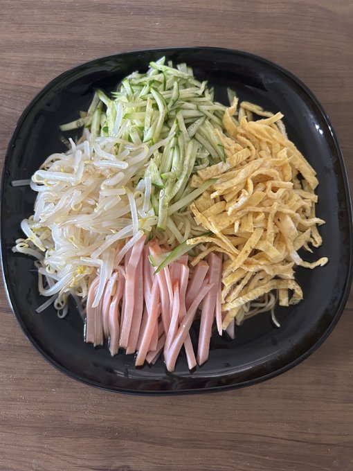 A top-down view of a black plate filled with a neatly arranged meal consisting of bean sprouts, shredded cucumber, sliced ham, and shredded omelet.