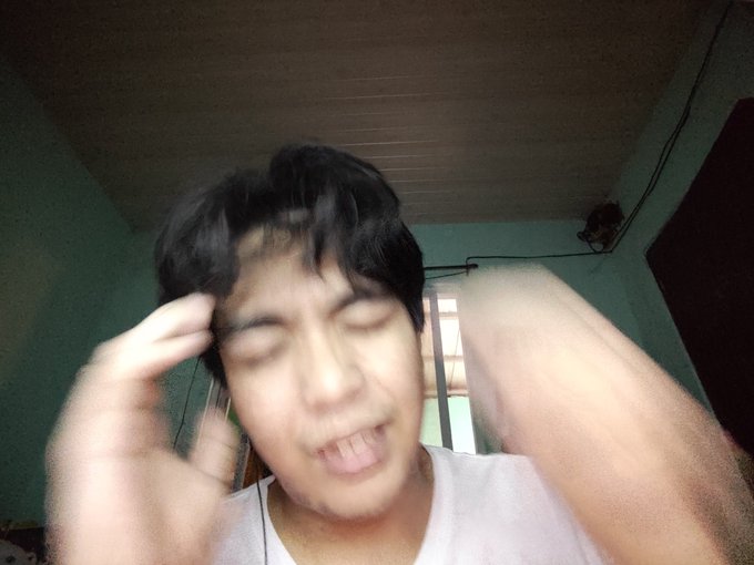 A blurry selfie of a young man with closed eyes and an open mouth, making a hand gesture near his head while wearing a white tank top in a room with green walls.