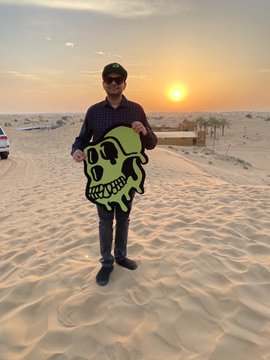 A young man wearing a cap and sunglasses stands in a sandy landscape at sunset, holding a green skull-shaped cutout.