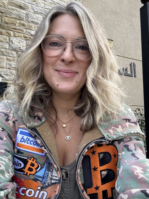 A blonde woman with glasses smiles outside a building while wearing a camouflage jacket covered in Bitcoin patches and a "Bitcoin" iron-on, paired with delicate eye- and heart-shaped necklaces, ready for day 4 of her shamanic counseling class.