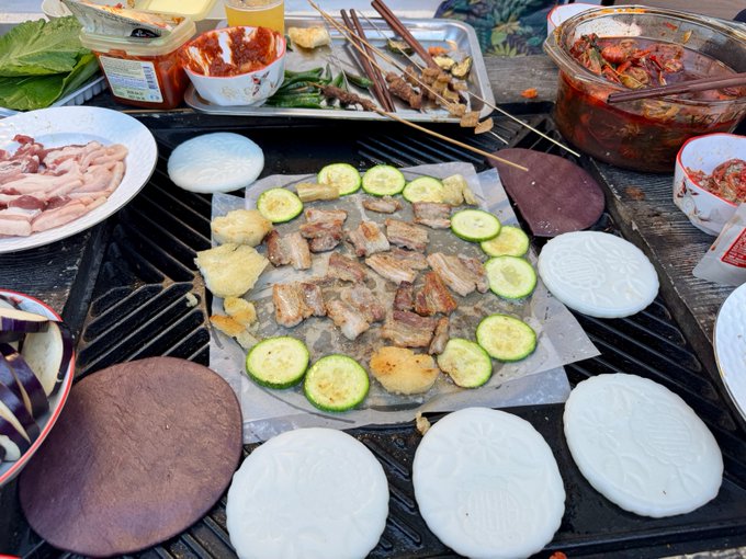 An outdoor grill is laden with various foods including sliced meat, zucchini, pineapple, skewers, and white steamed buns, surrounded by containers of kimchi and dipping sauces.
