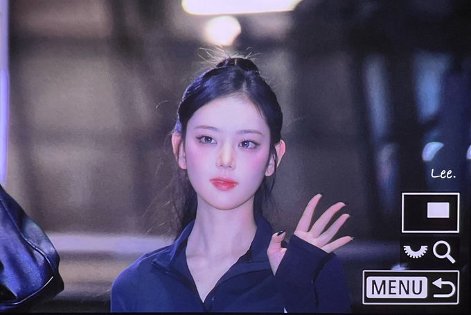 A young woman with dark hair styled in a half-updo, wearing a black top, posing with her hand raised; the image has a camera interface overlay with 'MENU', 'Lee.' text, and icons for eyes and a magnifying glass.