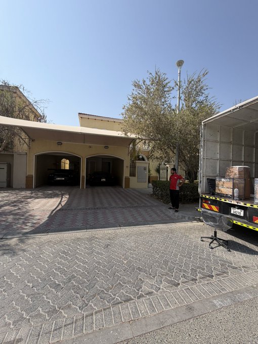 A moving truck is parked outside a residential garage complex, where a worker in a red shirt stands near the open back filled with cardboard boxes and furniture on a bright, sunny day, capturing the active process of relocating.