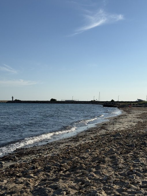 A daytime view of a pebbly beach meeting calm blue water, with a breakwater, a lighthouse, and street lamps visible in the distance under a bright blue sky.