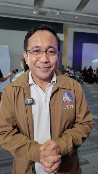 A man wearing a brown jacket with a "DICT" logo and a lavalier microphone smiles at the camera, identifying him as DICT Undersecretary for e-Government David Almirol during an interview at the Philippine Blockchain Week.