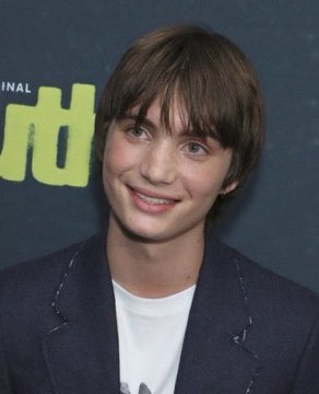 A young man with brown hair wearing a dark blazer over a white t-shirt, smiling in front of a green backdrop.