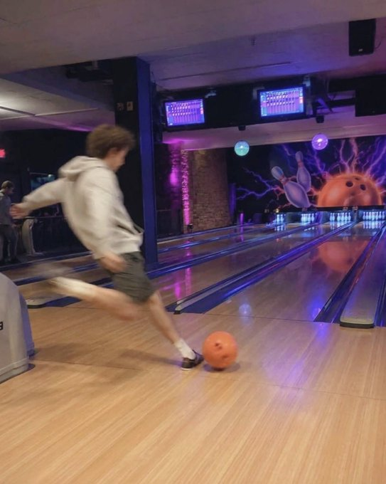 A young man in a white hoodie and shorts bowling at a bowling alley with neon purple lighting, releasing an orange bowling ball down the lane.