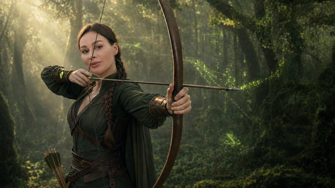 A woman with braided hair dressed in medieval-style archer attire, drawing back a bowstring with green magical sparkles trailing from the arrow, set in a sunlit forest.