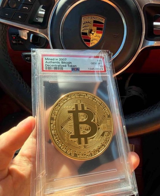 The image features a person holding up a gold coin, which appears to be a Bitcoin or another type of cryptocurrency. The coin has a gold-colored design with a black border, making it stand out against the background. The coin is placed inside a clear plastic case that protects and showcases its value. The context suggests that this could be an advertisement for a product related to cryptocurrencies or digital assets, possibly aimed at collectors or those interested in investing in Bitcoin or other virtual currency.