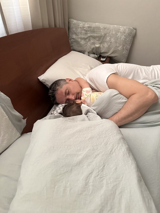A man peacefully naps in bed with a baby wearing a yellow floral outfit, embracing them tenderly. The cozy, intimate scene captures a quiet family moment—suggesting that despite the allure of new technologies, the simple joy of a parent's love and closeness with their child is truly unmatched.