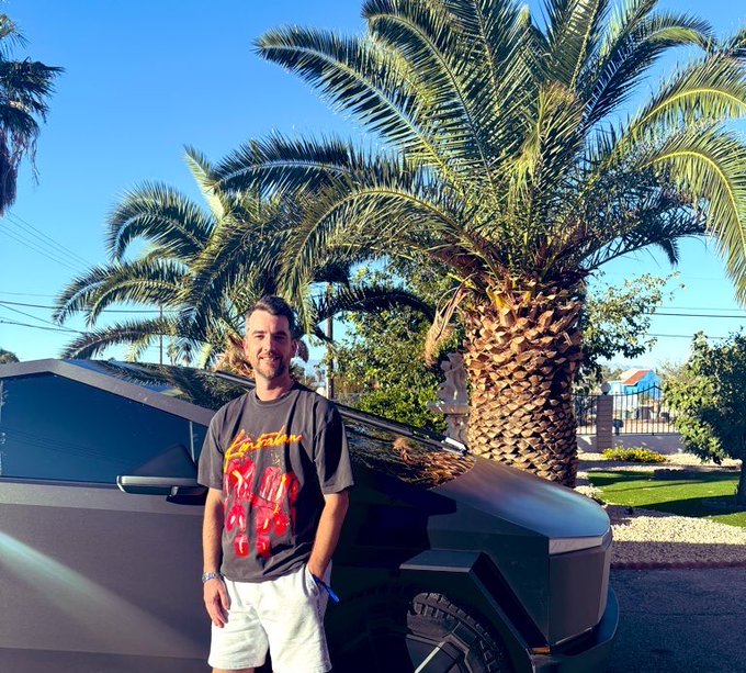 A man wearing a graphic t-shirt stands smiling in front of a Tesla Cybertruck and a palm tree.