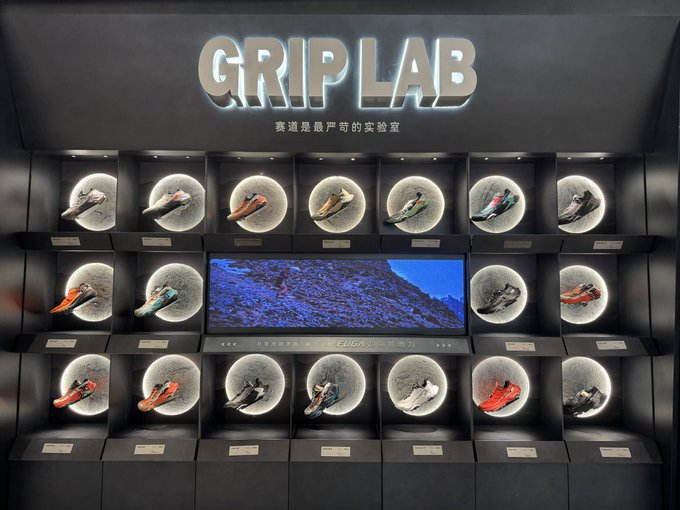 A retail display for 'GRIP LAB' showcasing multiple running shoes illuminated in circular compartments around a central screen showing a runner on a trail.
